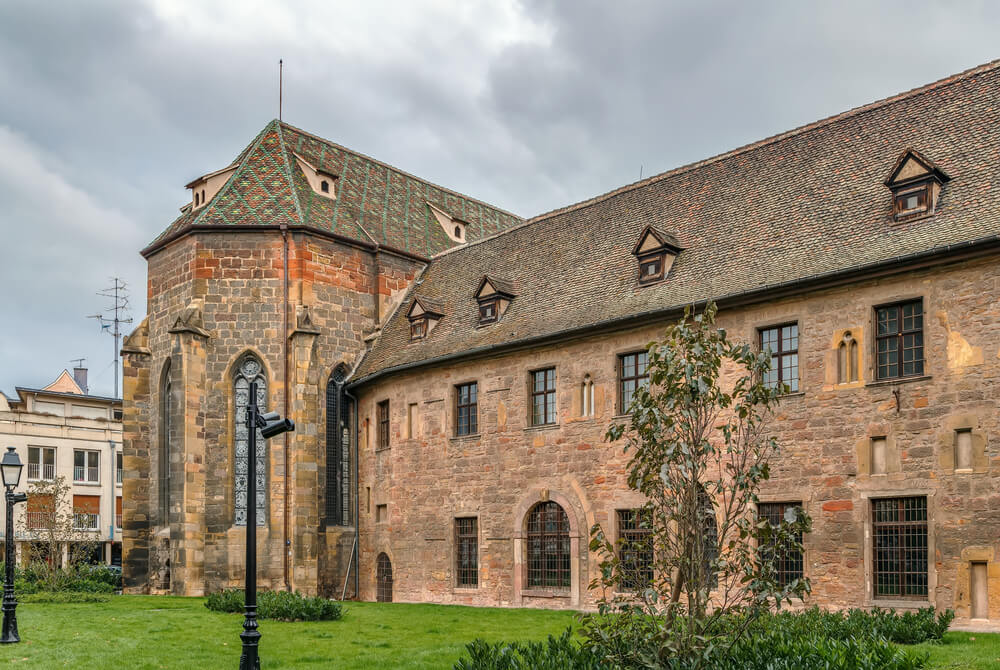 The Unterlinden Museum in Colmar emerges as a splendid blend of medieval architecture and modern surroundings. The building's defining feature is its green and red patterned tile roof, reminiscent of traditional Alsatian design. A sturdy, well-preserved tower, constructed from brown and beige bricks, anchors the corner, with Gothic windows