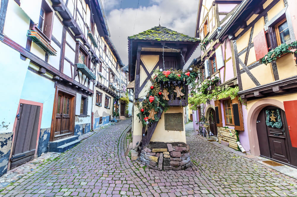 A charming cobblestone street in a European town, lined with half-timbered houses exhibiting a mix of colors like blues, pinks, and whites. The houses are adorned with festive decorations including wreaths, garlands, and Christmas ornaments. Each dwelling has unique architectural details like overhanging balconies, wooden shutters, and decorative moldings. A prominent feature is a covered passage with a conical, shingled roof, supported by stone pillars