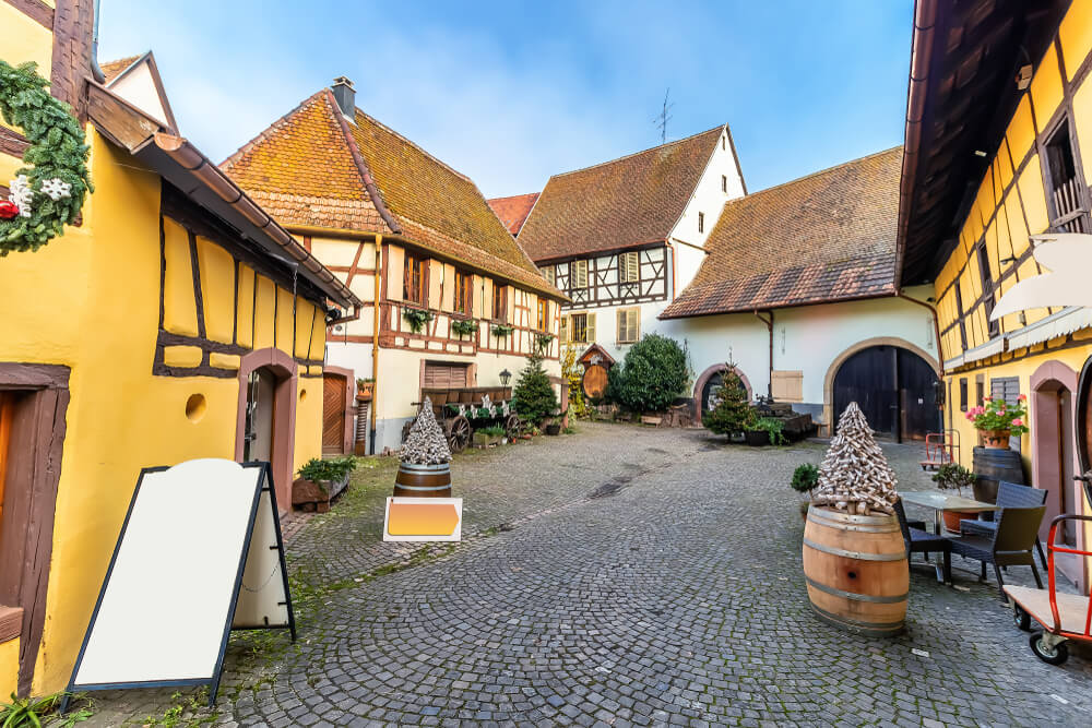 A quaint courtyard surrounded by traditional half-timbered houses with richly colored facades. Features include a cobblestone pavement, an empty white signboard, a decorative cone-shaped structure atop a wooden barrel, and seating arrangements. 