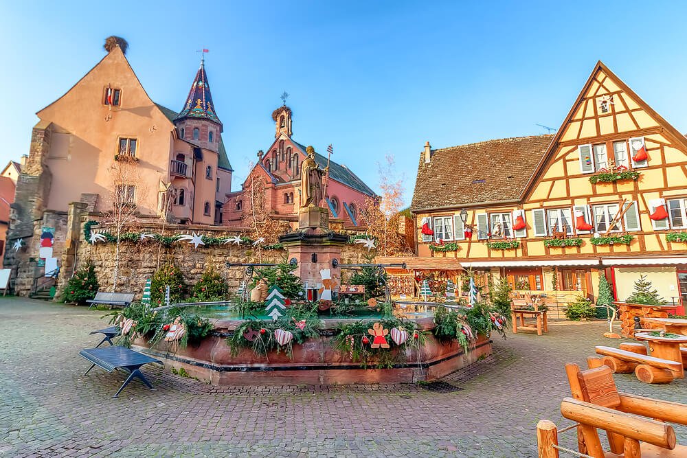 A picturesque town square adorned with festive decorations. Prominent features include a historical fountain surrounded by greenery and holiday ornaments, traditional half-timbered buildings with vibrant facades, and a church with intricate architectural details. The atmosphere is imbued with the spirit of the holiday season, complemented by benches, wooden setups, and hanging Christmas stockings on building windows.