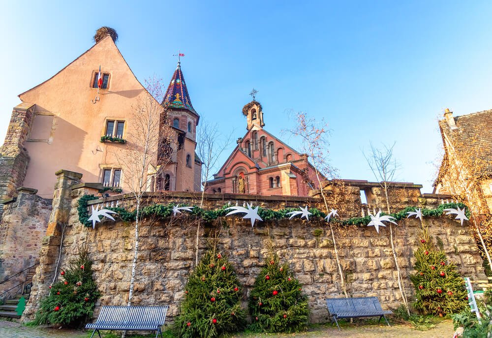 A picturesque European town square during the daytime, showcasing historic and architecturally distinct buildings. To the left, a peach-colored building with a whimsically pointed roof and a small balcony contrasts with the gothic spire of a tower behind it. Adjacent to these, a majestic red-brick church with ornate details and a prominent bell tower rises against the clear sky. A stone wall, adorned with white star decorations and flanked by tall evergreen trees with red ornaments, forms the foreground.