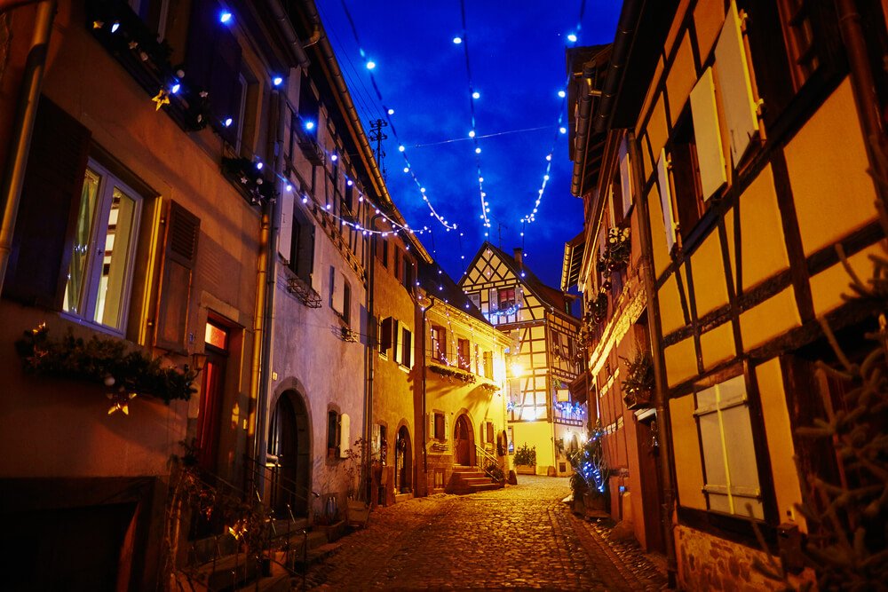 A magical nighttime scene in a European alleyway, illuminated by twinkling blue lights strung overhead. Traditional buildings, with features such as wooden beams and rustic stonework, line the cobbled street. The facades of the buildings are decorated with festive garlands, wreaths, and ambient lighting from windows