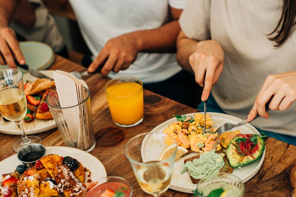 A close-up view of a dining table where two people are enjoying a gourmet brunch. In the foreground, there's a plate of French toast garnished with fresh berries and dusted with powdered sugar, accompanied by a side of syrup. Next to it, another plate showcases a vibrant assortment of foods: creamy scrambled eggs sprinkled with herbs, avocado slices garnished with radish and greens, and a slice of toast spread with a green dip