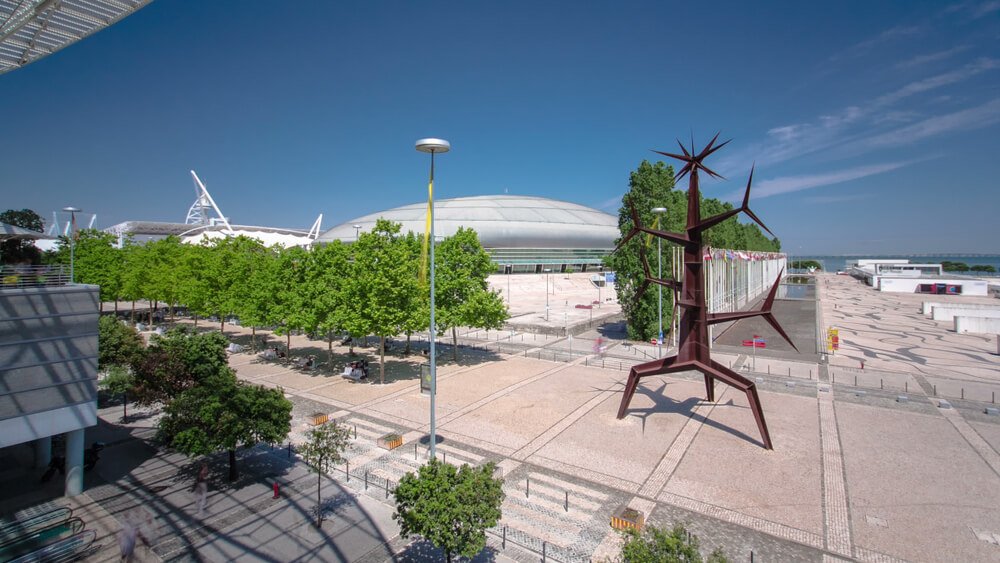 Modern urban plaza under a clear blue sky, featuring a row of neatly planted trees and a paved open space. In the foreground stands a large, abstract metal sculpture with spiky extensions. Behind the plaza, there's a distinctive building with a rounded, dome-like roof