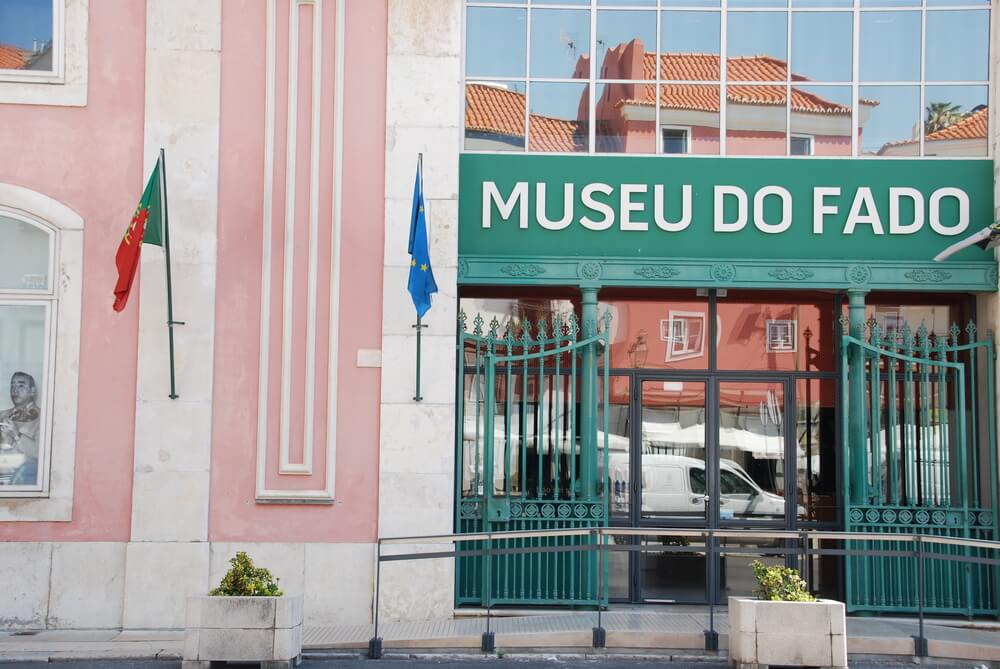 Facade of "Museu do Fado" with a green signboard, flanked by the Portuguese national flag and the European Union flag. The entrance features large glass windows and green ornate gates. The building has a pastel pink colored exterior and green sign.