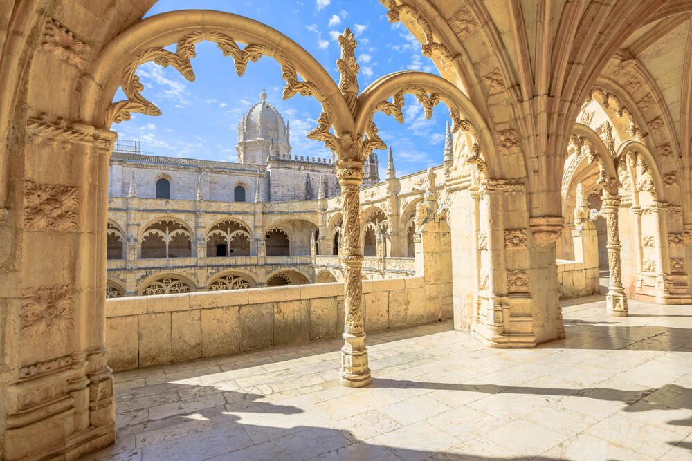 A sunlit Gothic courtyard with intricately carved arches and stone columns, overlooking a historic building, at the famed Jeronimos Monastery in Lisbon, a UNESCO site.