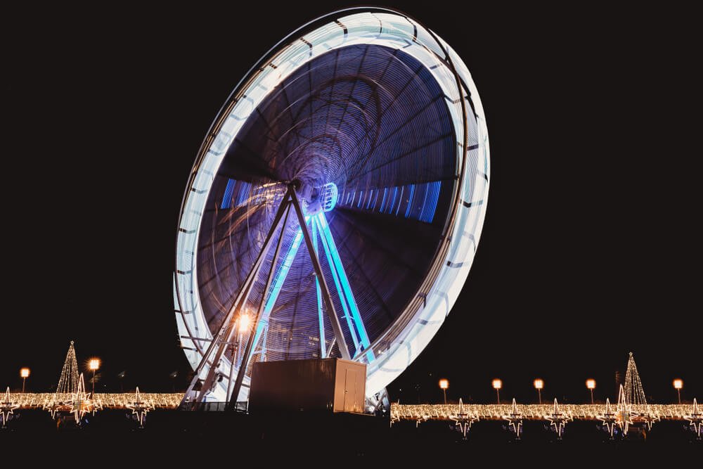 A luminescent Ferris wheel in motion at nighttime, its radiant blue and white lights creating a dazzling circular blur against a dark sky, with a background of illuminated star-shaped decorations and glowing lampposts at a CHristmas market in Lisbon in December