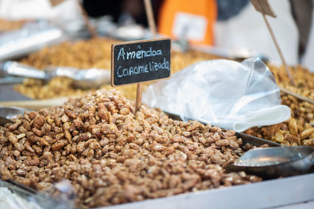 A close-up view of a tray filled with caramelized almonds, labeled "Amêndoa Caramelizada" on a chalkboard sign. Beside it are other trays of assorted treats, a metal scoop, and a clear plastic bag for packaging. The glistening almonds appear crunchy and sweet at a Christmas market.