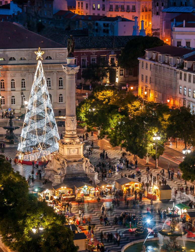 A festive city square at night with a luminous Christmas tree, a prominent statue on a column, bustling market stalls, and people milling about on a patterned pavement. Surrounding buildings emit a warm glow, adding to the ambiance.  