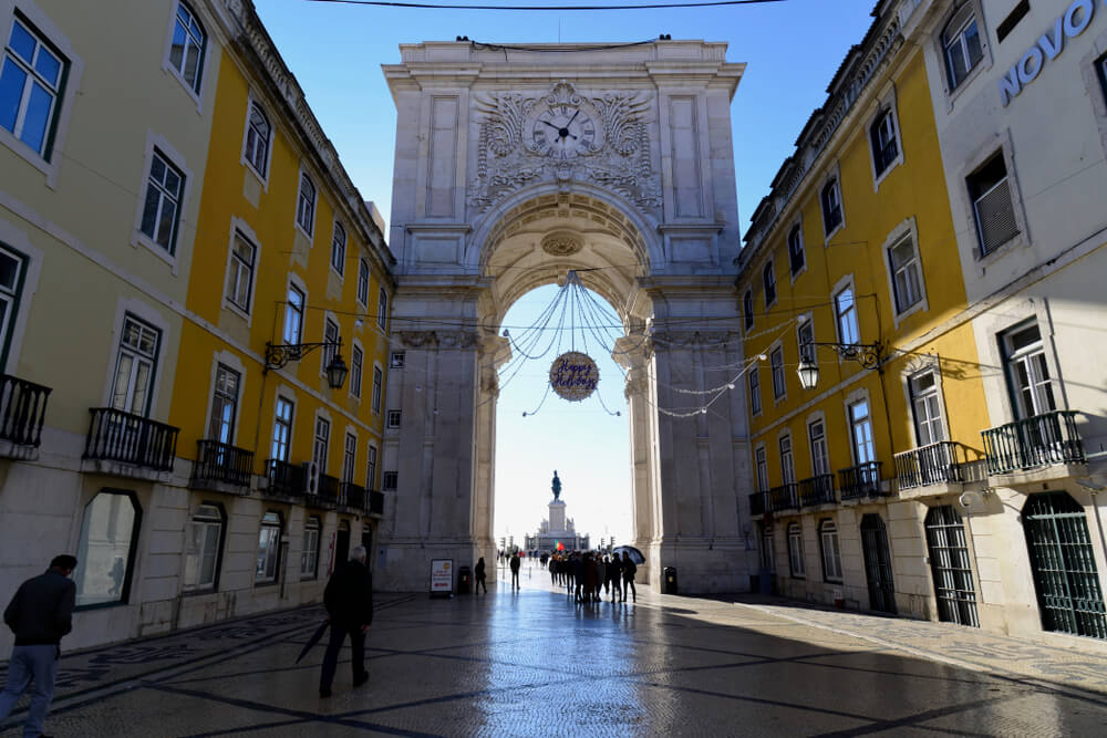 A picturesque urban scene of a grand archway with ornate stone carvings and a clock. Adjacent to the archway are bright yellow-colored buildings with white trims and Christmas decorations visible but not illuminated as it is still the daytime.