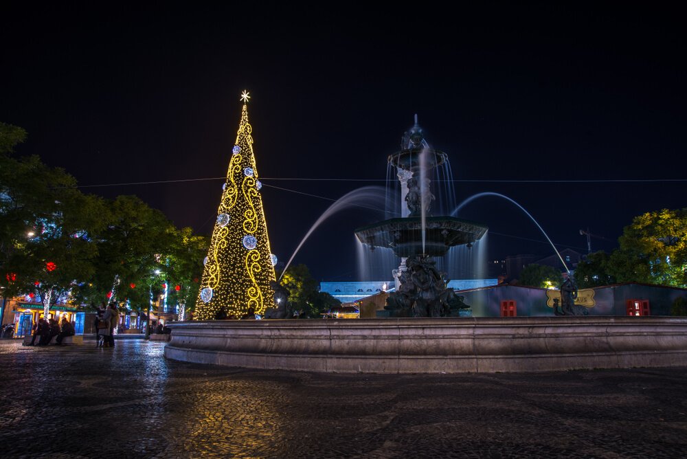 A vibrant city square at night featuring a towering illuminated Christmas tree adorned with golden lights and swirling designs. Beside it stands an elegant fountain with water gracefully arching from its tiers, silhouetted against the ambient city lights. People gather and relax in the foreground, while festive decorations and cityscape details in the background.