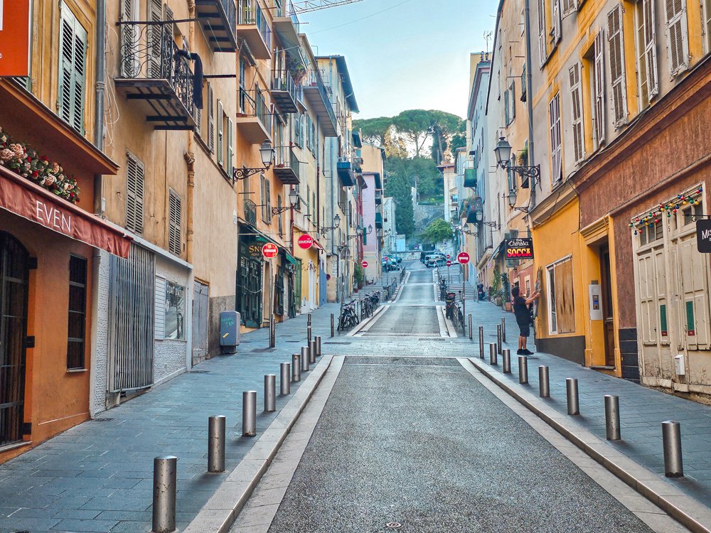 An empty street in the old town of Nice looking up to the view of Castle Hill on the corner, a sign for a socca restaurant, and a person getting the buildings ready for the day.