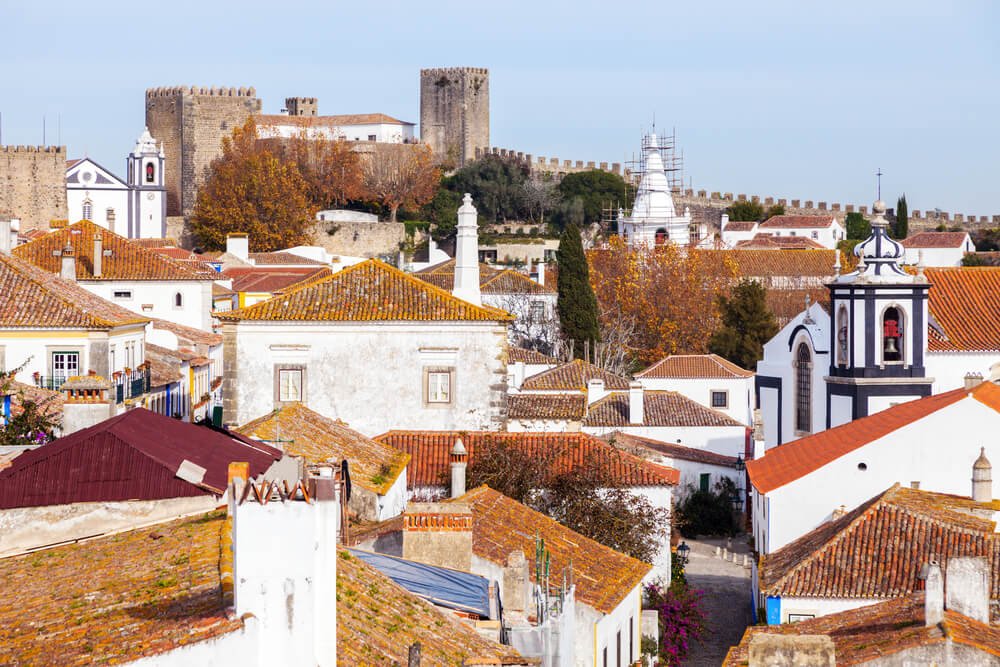 A picturesque townscape with orange-tiled rooftops, white buildings, and a prominent medieval castle in the background, with a white CHristmas tree like structure being built for the holidays, and some festive decorations.