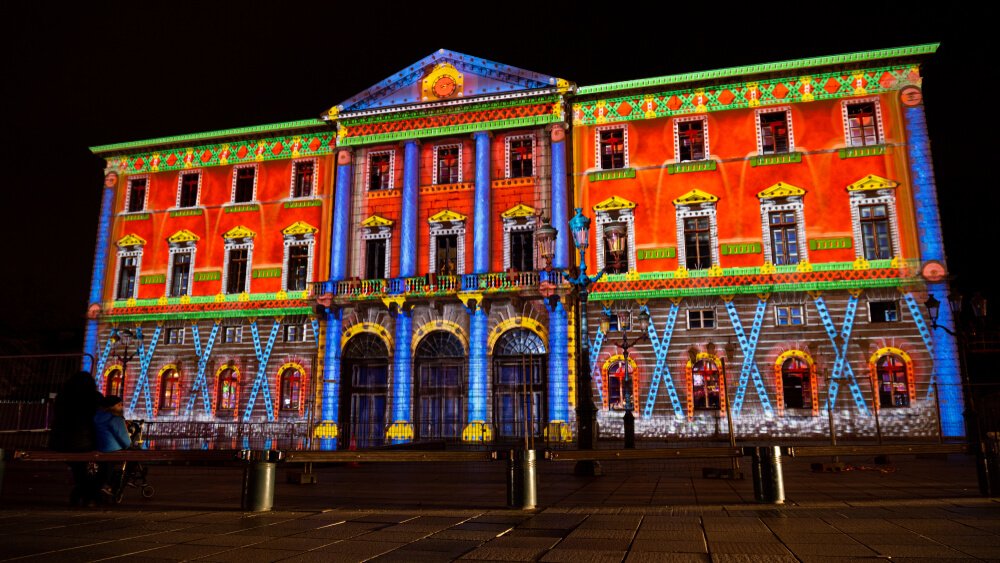 A vibrant and colorful light projection on a grand historic building during nighttime. The projection displays intricate patterns and designs, transforming the façade into a tapestry of vivid hues, primarily in red, blue, and green