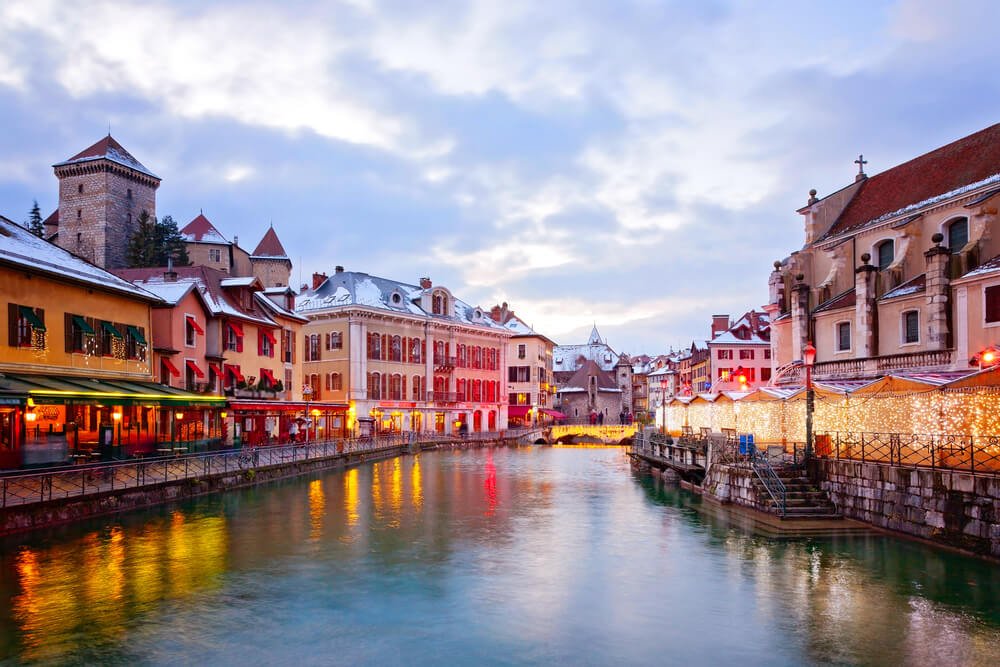 A picturesque European town scene during twilight. The canal waters calmly reflect the warm glow of lights from the surrounding buildings. Historic structures, including a stone tower and various architectural styles, line the canal. These buildings are adorned with festive lights and decorations, adding to the ambiance. 