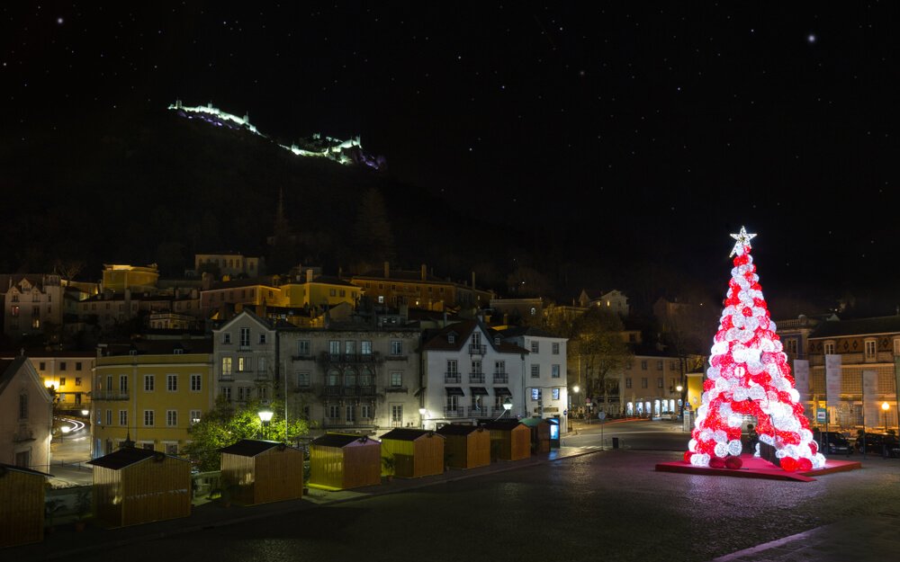 A picturesque nighttime scene of a Sintra square during the holiday season. Illuminated buildings line the streets, A vibrant Christmas tree, adorned with radiant red and white lights, and lights up on the hills above the town.