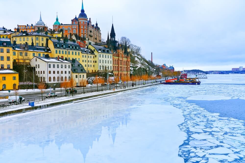Waterside perspective of a winter scene in Stockholm with castle, royal palace, yellow buildings, boat, and icy waters.