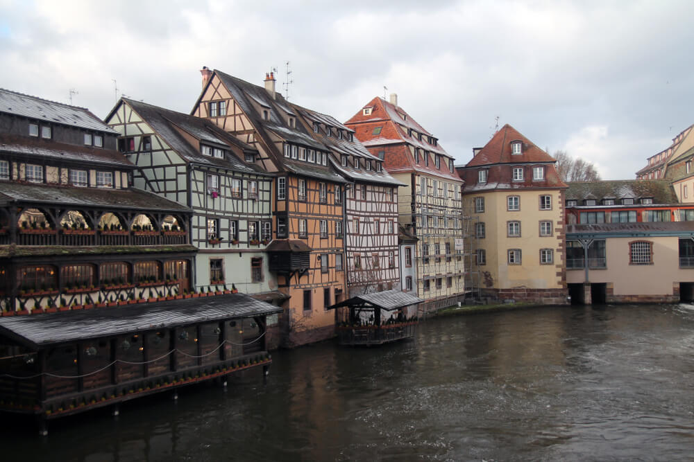 Colorful historic half-timbered buildings along a river, with a covered wooden terrace on the water's edge while on a boat cruise