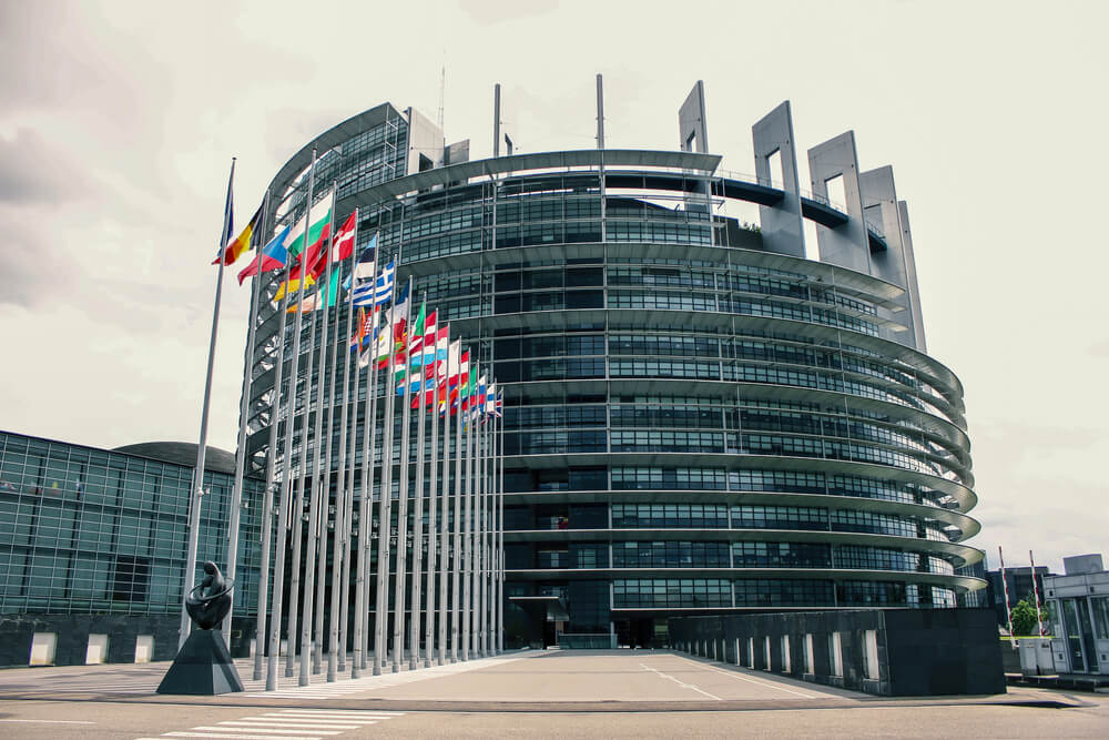 Modern multi-layered glass building of the European Parliament in Strasbourg, with a series of national flags on poles displayed in the forefront, under a cloudy sky in Strasbourg in winter.