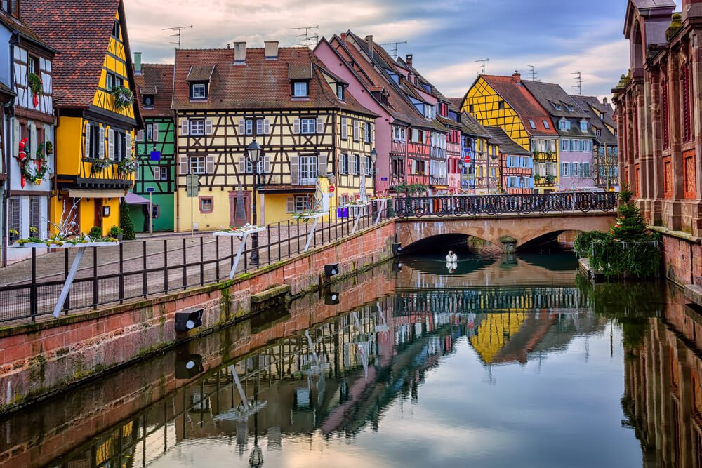 Picturesque canal scene with vibrant half-timbered buildings reflecting on calm water, a stone bridge in the background, and festive decorations adorning the facades in the town of Colmar in winter.