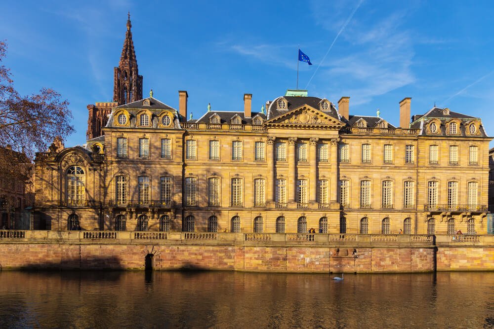 A grand European building with ornate architecture stands beside a canal, with a Gothic church tower in the background, under a clear blue sky.