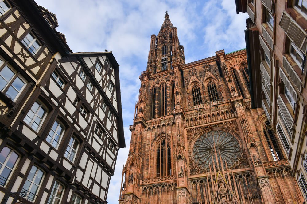 Street view in Strasbourg featuring traditional half-timbered houses juxtaposed against the intricate Gothic architecture of Strasbourg Cathedral with its ornate facade and towering spire against a blue sky.