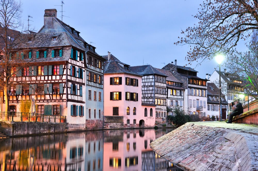 Tranquil dusk scene by a canal, showcasing a row of charming half-timbered buildings reflecting on the water, with a few people seated on the cobblestone bank under the soft glow of a streetlamp.