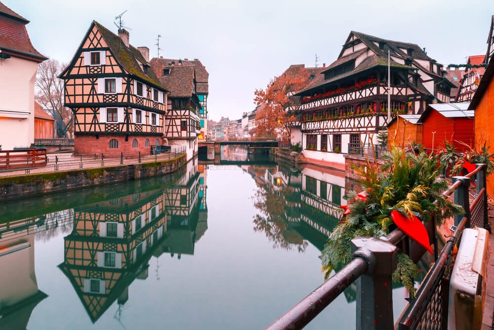 Historic half-timbered houses along a calm canal, reflecting perfectly in the water, with a decorative railing adorned with festive greenery and red bows in the foreground, set against a soft gray cloudy sky.