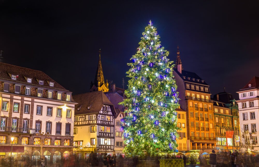 A brightly illuminated Christmas tree stands tall in a historic town square during nighttime, surrounded by traditional European buildings, with a church spire in the background. The tree's shimmering lights contrast beautifully with the warm glow emanating from the windows of the buildings, while blurred figures of people indicate activity and festivity in the square.