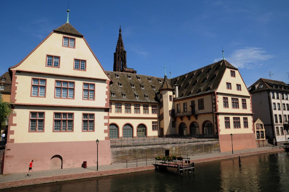 A picturesque European town scene featuring traditional buildings with pastel-colored facades and steep pitched roofs, situated alongside a calm canal. In the background, a Gothic-style church tower rises, contrasting with the more modest structures. A person in red is captured mid-motion while jogging.