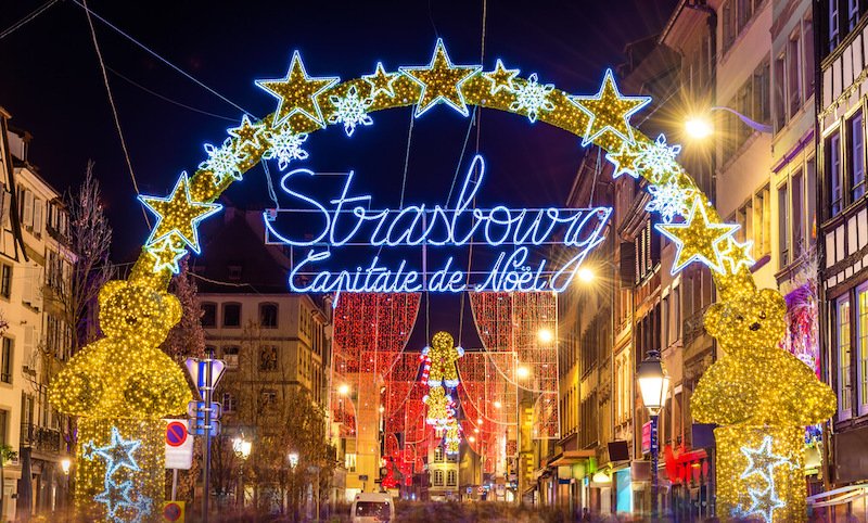 Nighttime view of Strasbourg's festive streets adorned with shimmering star-shaped lights, glowing gold bear decorations, and illuminated text reading 'Strasbourg Capitale de Noël' (Strasbourg, Capital of Christmas) against a backdrop of historic buildings.