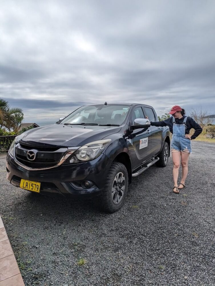 Allison Green wearing a hat, sweater, and overalls, standing next to a large pickup truck while visiting Fiji