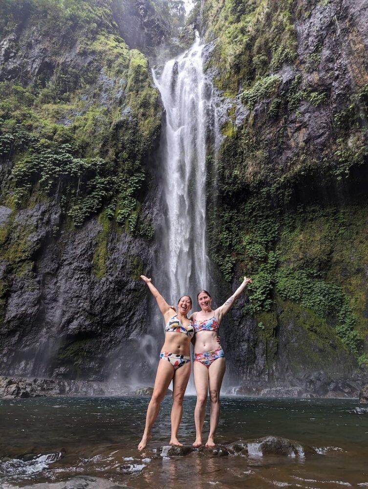 Allison and Deanne standing in front of the waterfall with their arms up enjoying the waterfall and showing its scale