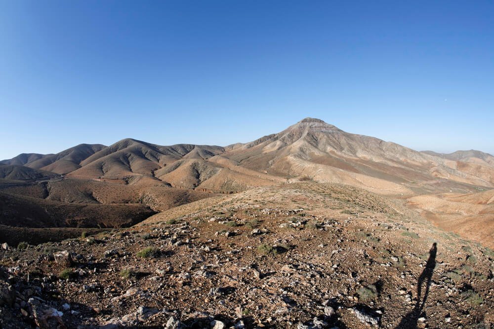 A person's shadow showing who is taking the photo in a volcanic landscape with barren peaks on a sunny cloudless day in Fuerteventura hiking