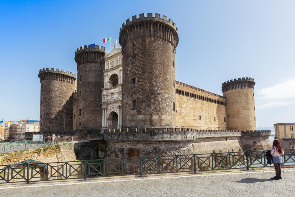 Woman in winter weather clothing standing in front of the castle Nuovo in Naples, with an italian flag flying, an indoor and outdoor castle complex great to visit in colder months