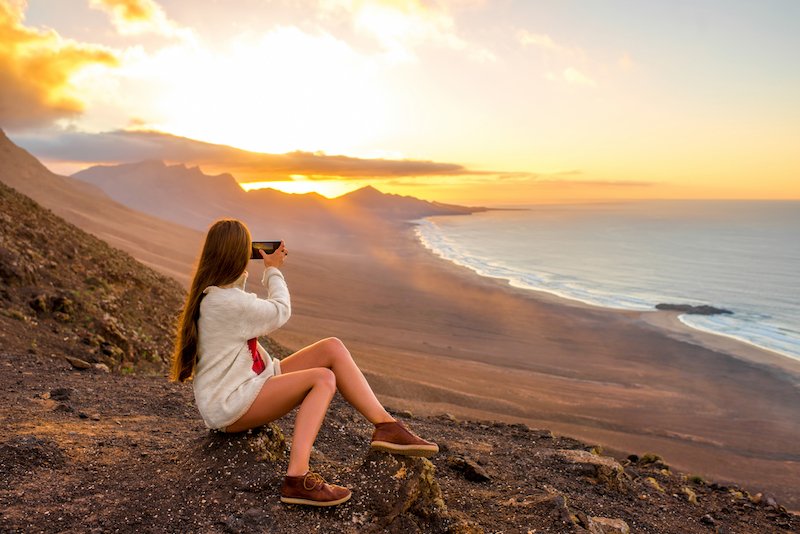 Young woman photographing with smart phone beautiful landscape with beach and mountains on the sunset over the Cofete Beach in Fuerteventura Spain