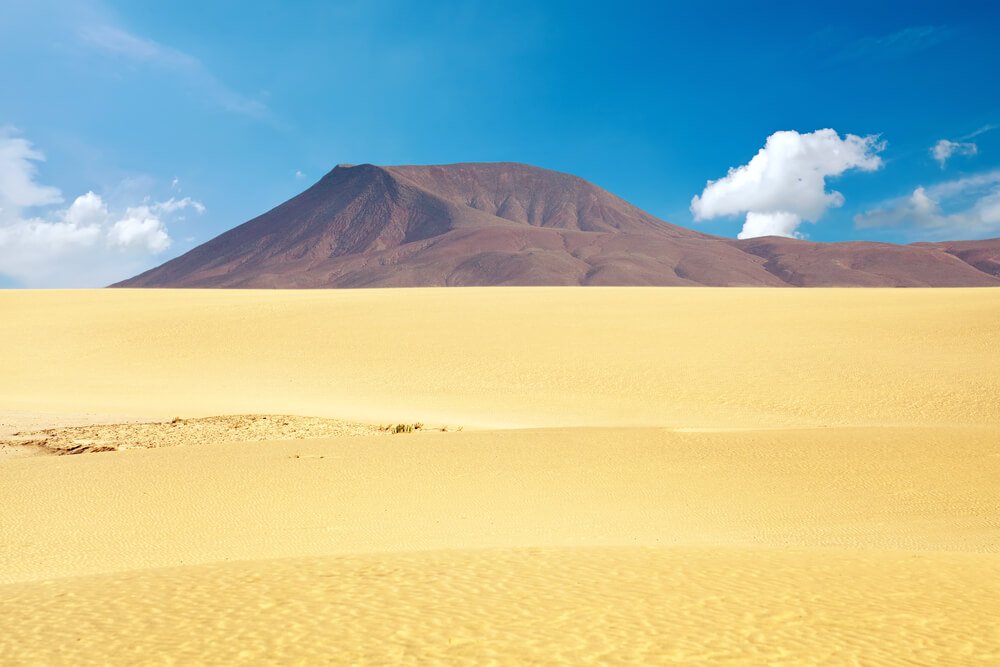 yellow sand dunes in corralejo in fuerteventura with a reddish mountain volcano cone in the distance, evoking a unique contrast of landscape