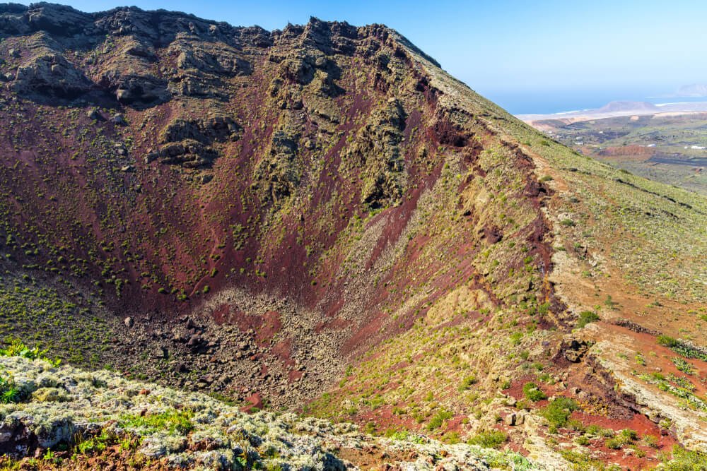 Red volcanic soil of volcano Corona with greenery and red dirt and view of the water in the background