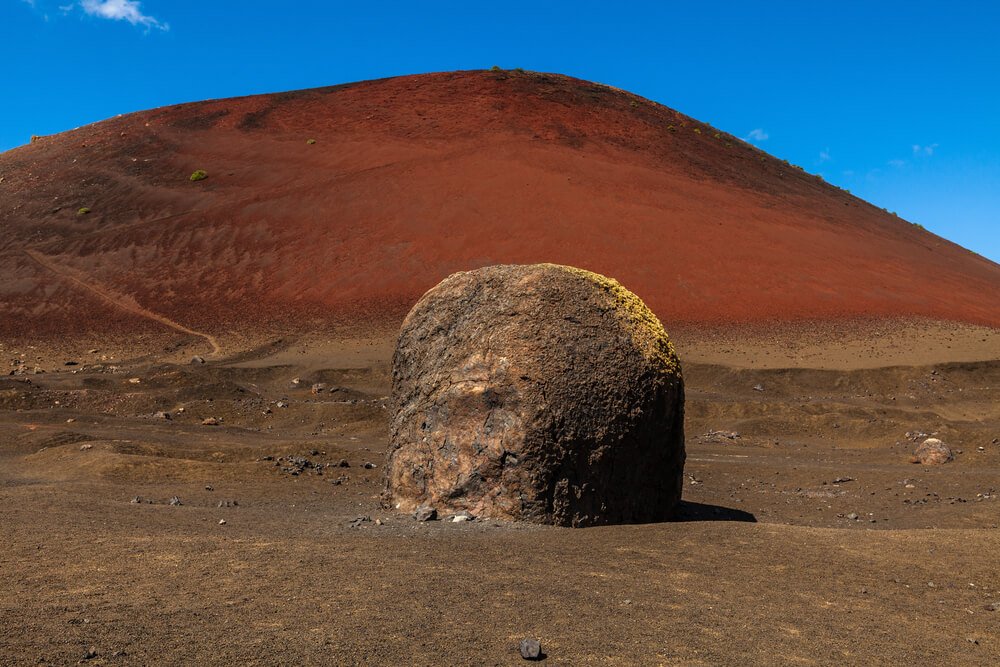 Volcanic bomb in front of volcano Montana Colorada in Lanzarote with reddish dirt of the volcano behind it and clear blue sky on a day hiking through Lanzarote