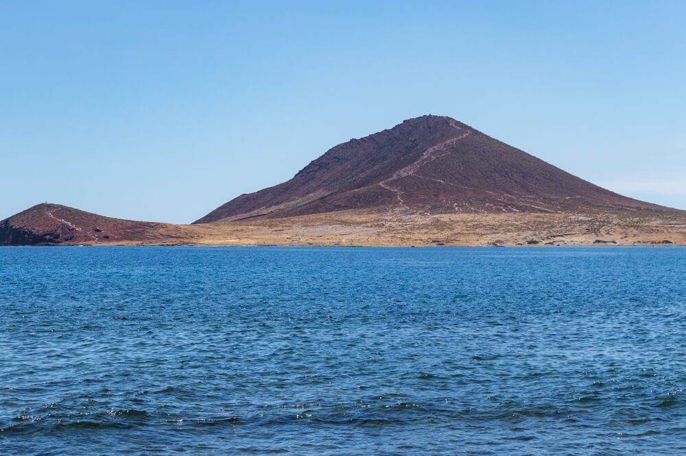 View of the Montana Roja or red mountain from across the sea with beautiful calm ocean water and hiking trail to the summit of the mountain visible even from a far distance
