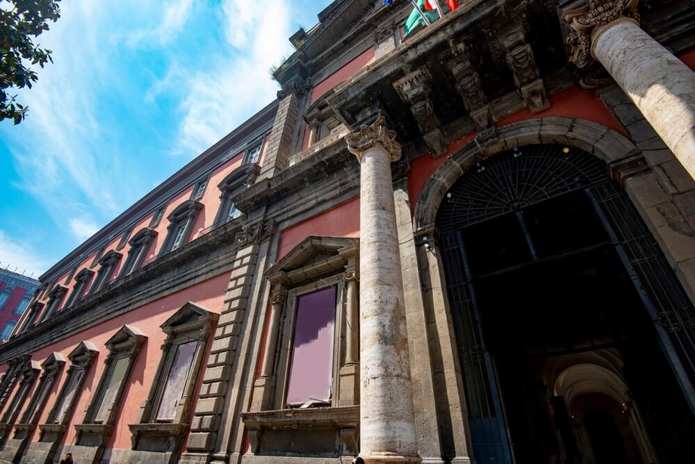 building entrance to the Naples archaeology museum in a salmon pink ornate building with italian flag on top of it