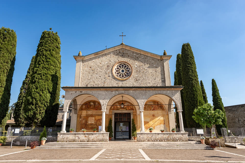 The front facade of a classic stone church, the Sanctuary of Madonna del Frassino, with a large circular window and an arched entrance, flanked by tall, narrow cypress trees against a clear blue sky. 