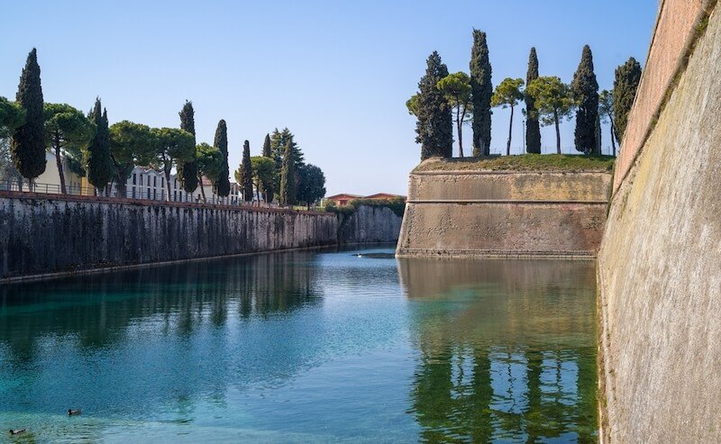 A serene water channel flanked by tall, fortified stone walls and lined with cypress trees under a clear blue sky. The tranquility of the scene is accentuated by the reflection of the trees in the calm water while visiting Peschiera del Garda town, a UNESCO site.