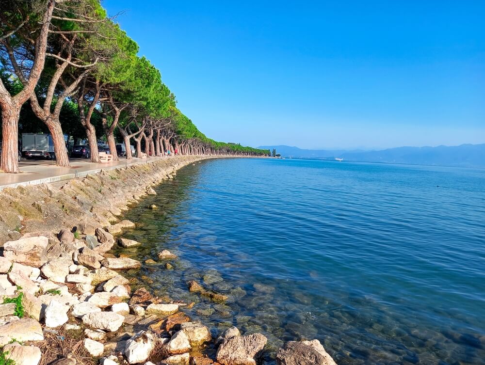 Lakeside promenade where a row of tall trees lines a paved walkway, following the gentle curve of a clear blue waters of Lake Garda, with a rocky shore.