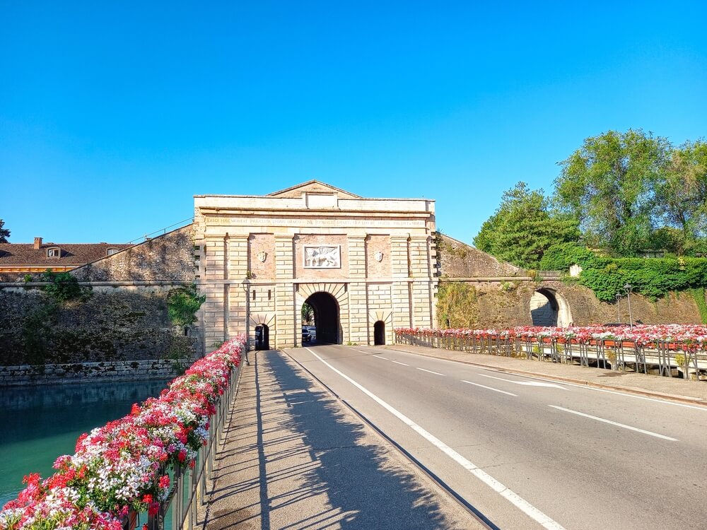 An image of a historical stone gateway leading to a fortified structure, with a road passing through it, adorned with vibrant flowers along the balustrade next to a waterway under a clear blue sky.