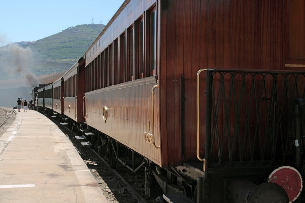 The Douro Valley historical train with traditional wooden compartments of the train and powered by steam which you can see at the back of the train, people walking away from the train and hills and vineyards in the distance