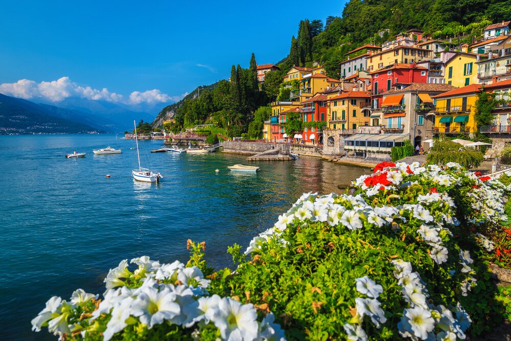 White flowers in flower boxes on the edge of a passageway through Lake Como's lakeside walkways with a view to the colorful red, orange and yellow houses