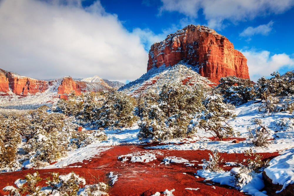 A snow-covered red rock landscape in the winter in Sedona with its characteristic geography covered in trees and snow with blue sky and clouds behind it
