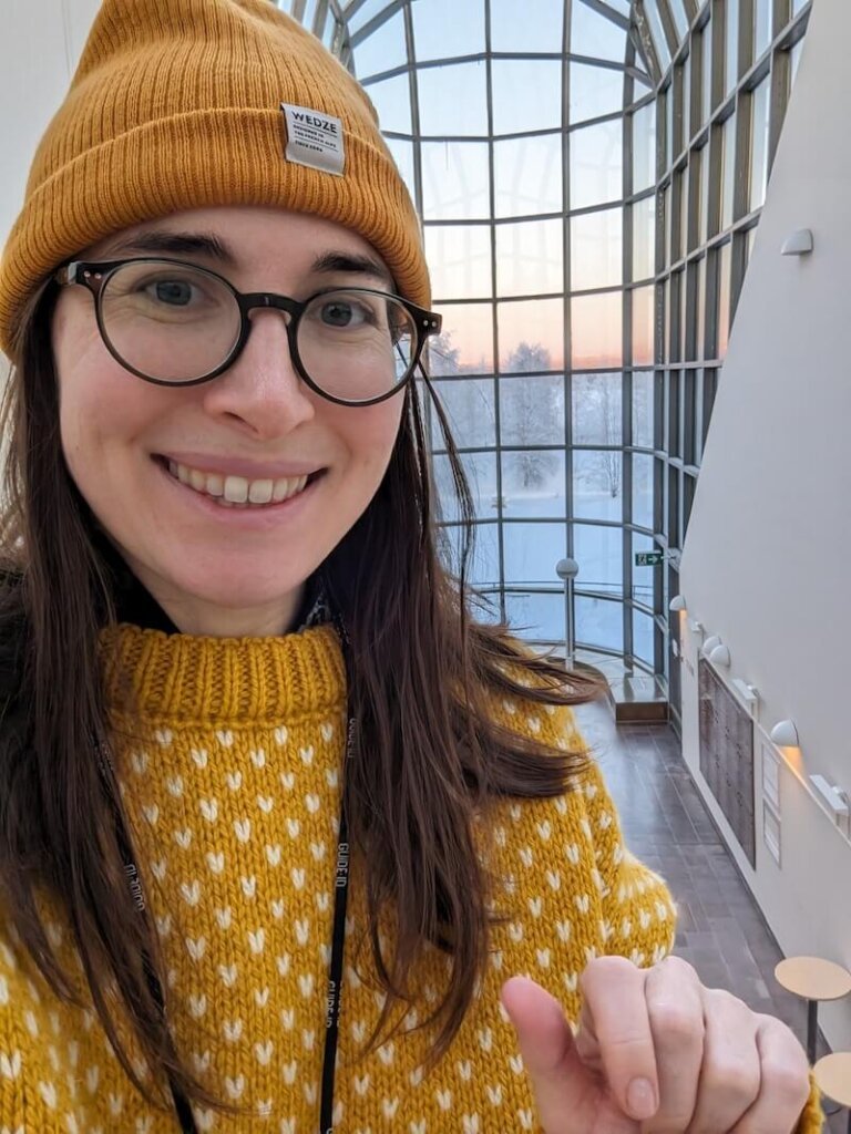 Allison Green, the author of the article, wearing a yellow sweater and hat, smiling at the camera with the building's unique architecture visible behind her