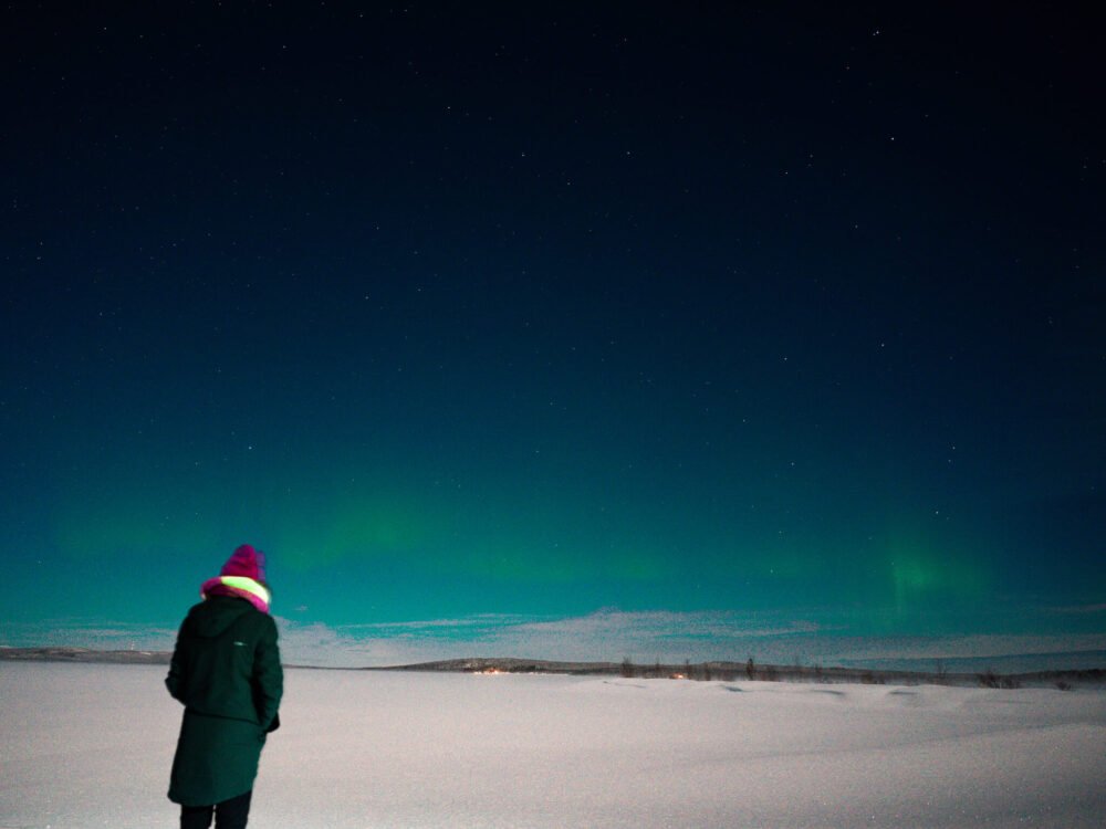 Allison Green standing in front of the Northern lights on a frozen lake in Finland with her back to the camera